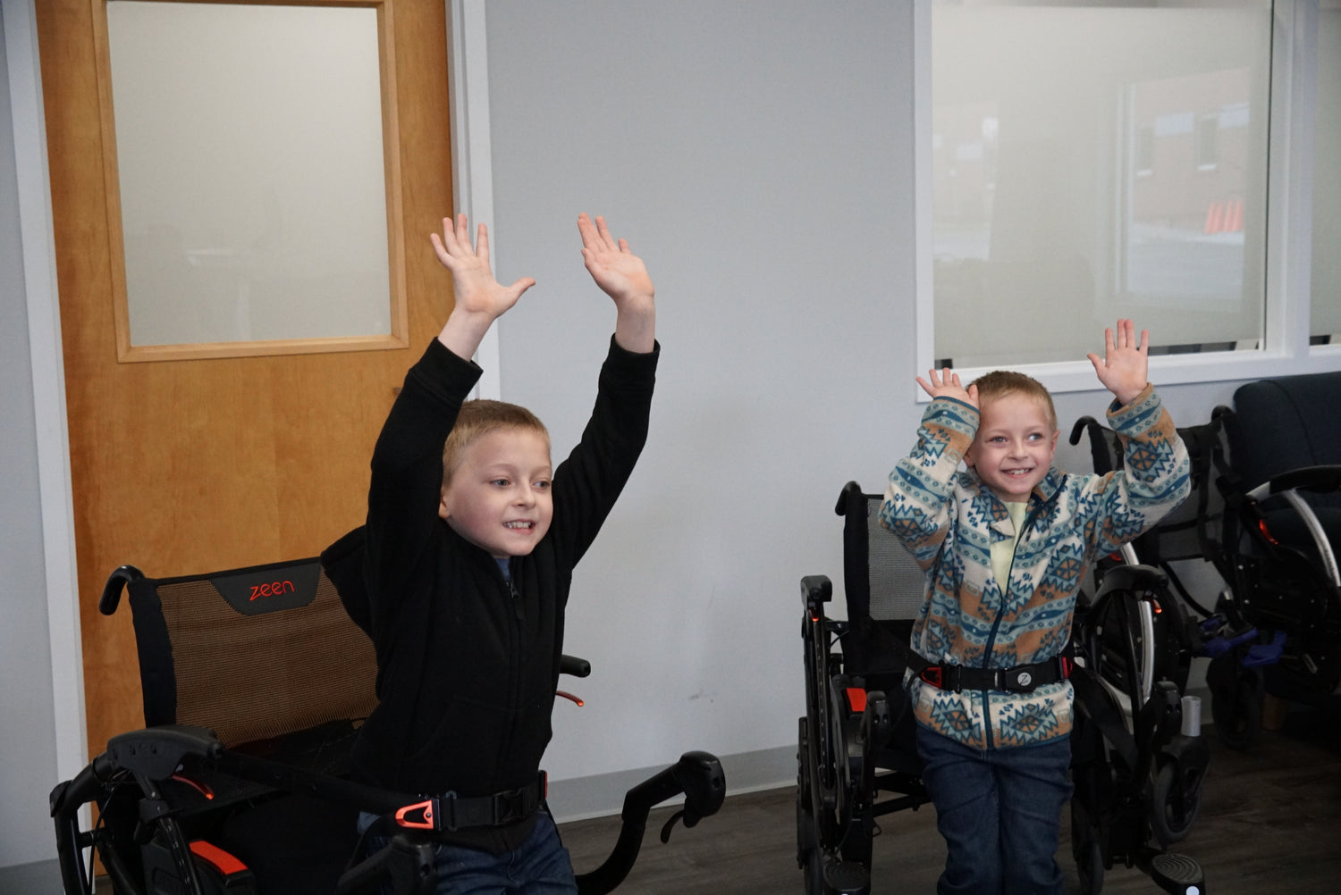 Two children standing in Zeen sit stand walkers indoors, smiling with both hands raised; waist belts fastened for support and black mesh-backed frames visible.