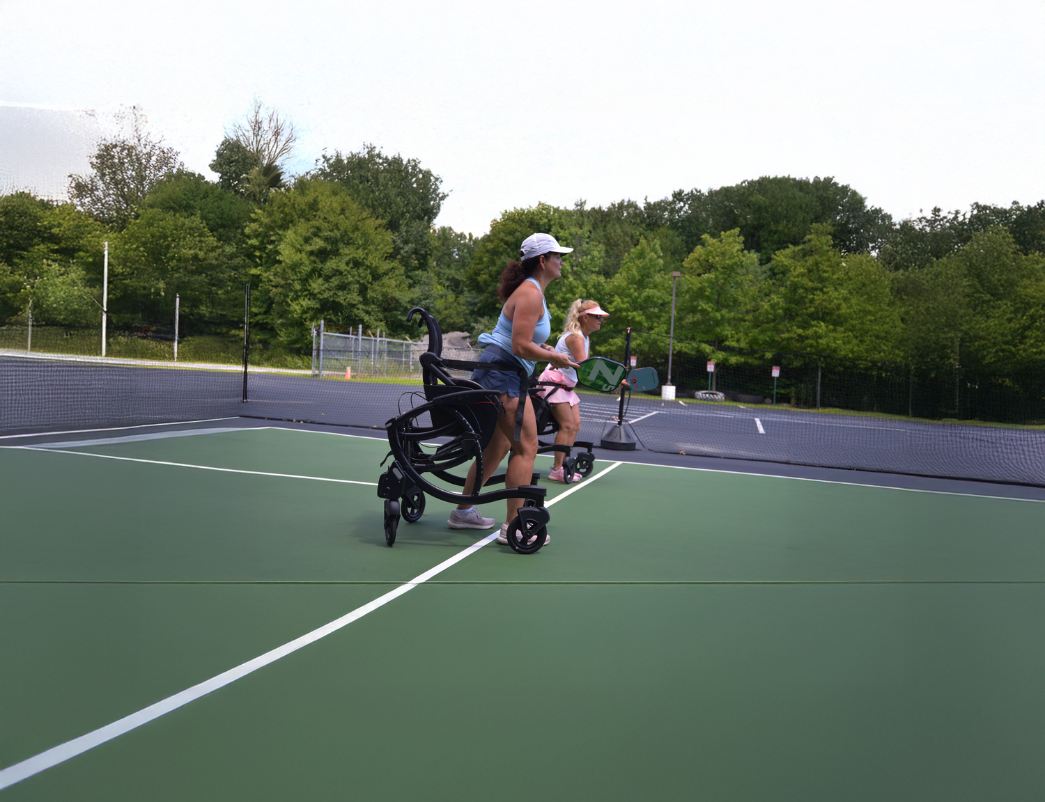 Two women on an outdoor pickleball court, one using a Zeen sit stand walker while holding a paddle, the other standing beside her ready to play.