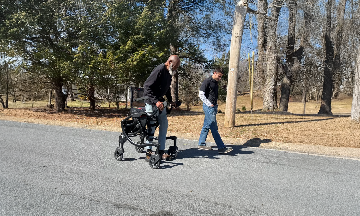 Person using a Zeen sit stand walker moves up a neighborhood street while another person walks beside them; four wheeled frame with integrated seat and hand brakes visible.